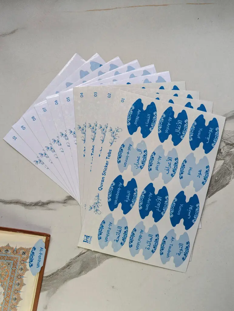 Quran sticker tabs by Iman & Ink Store, sheets of light and dark blue surah tabs fanned out, with an Al-Fatihah tab on a Quran page